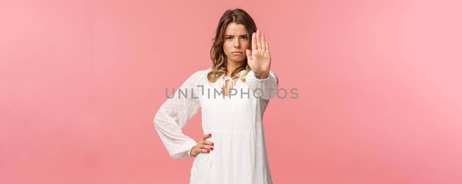 Waist-up portrait of serious-looking assertive and confident young blond female in white dress, extend arm in stop motion, frowning, demand quit, prohibit something, warning express disagreement.
