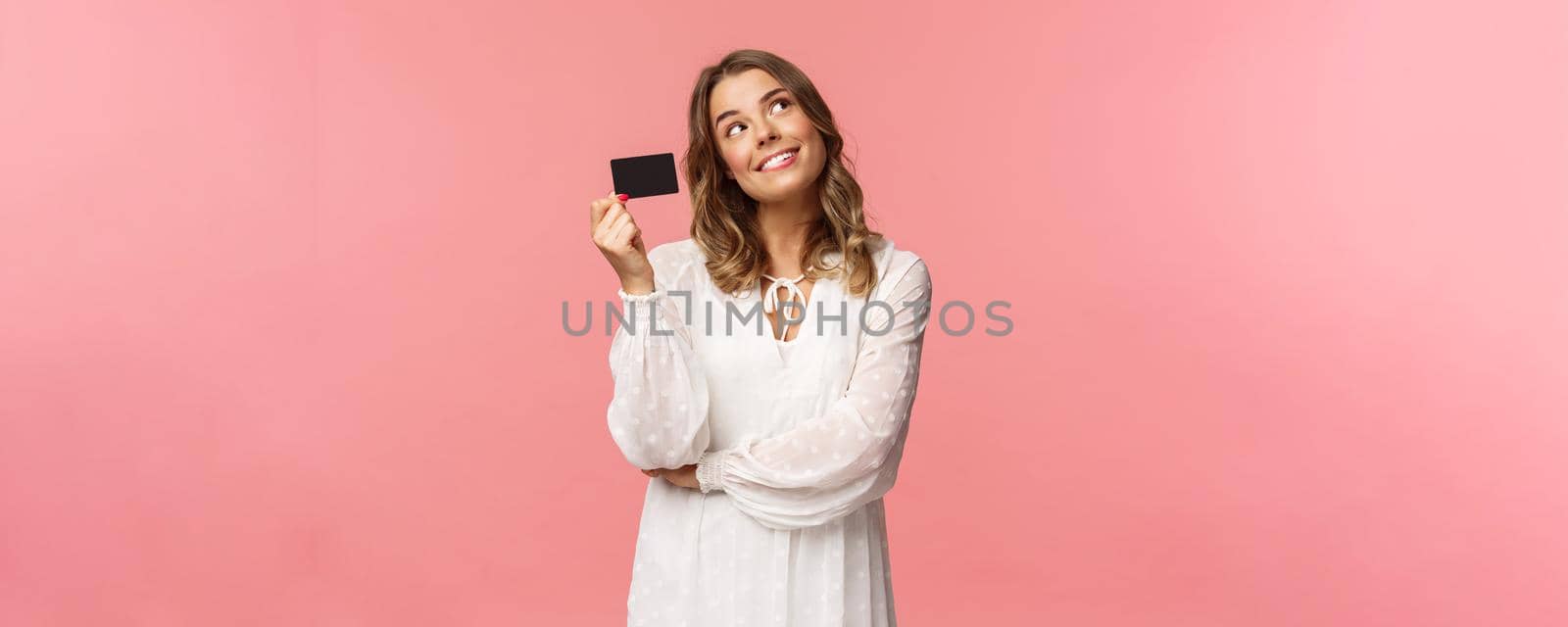 Portrait of cute tender feminine blond girl daydreaming about how she will waste her money on online shopping, look up dreamy, imaging as waiting delivery, hold credit card, pink background by Benzoix
