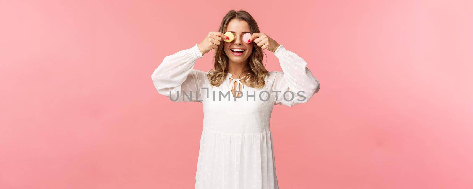 Holidays, spring and party concept. Portrait of carefree funny and cute blond girl in white dress, holding two macarons on eyes and smiling happy, like eating tasty desserts, pink background by Benzoix