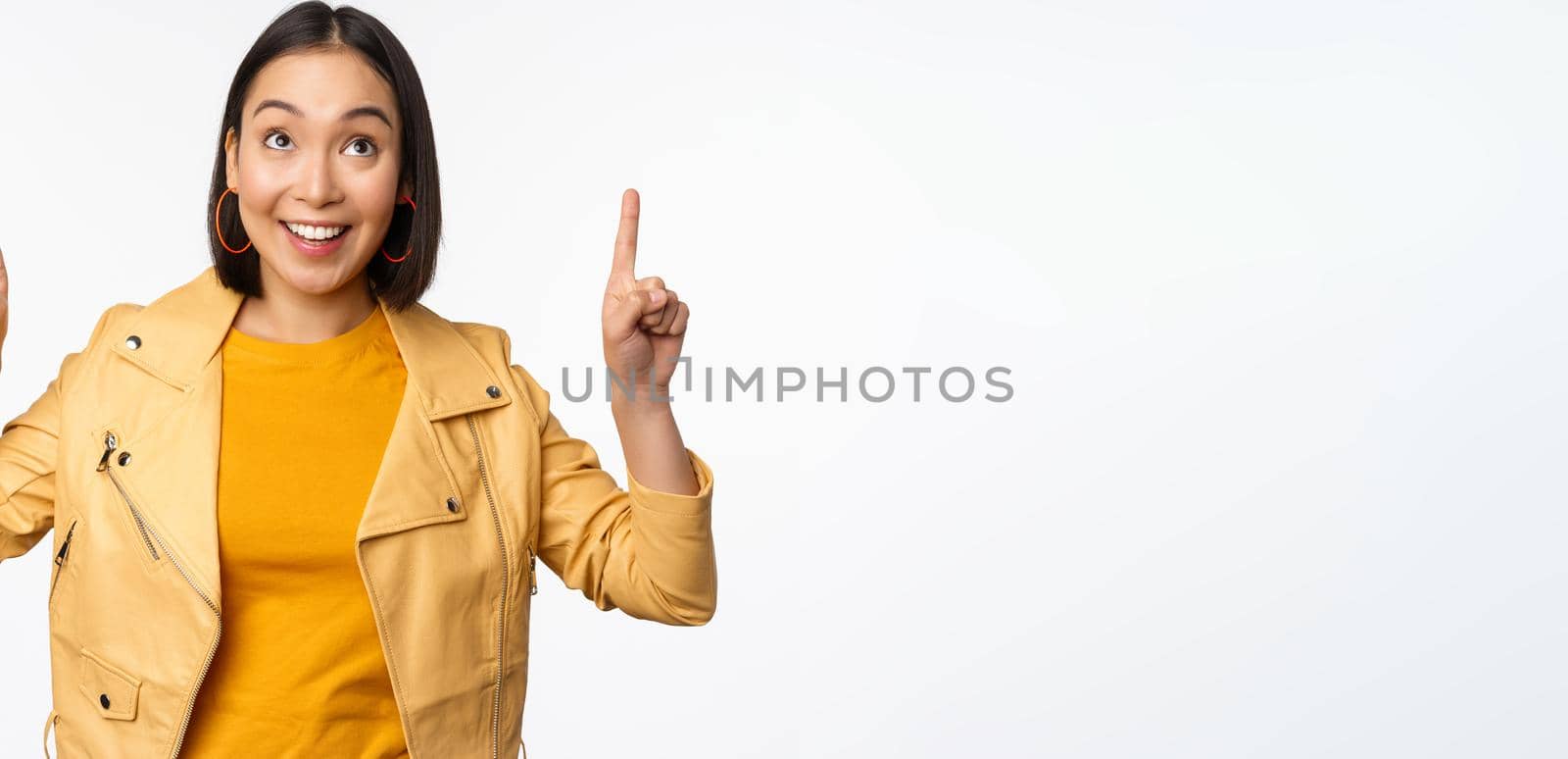 Image of smiling asian brunette woman pointing fingers up, showing advertisement with happy face, posing against white background.