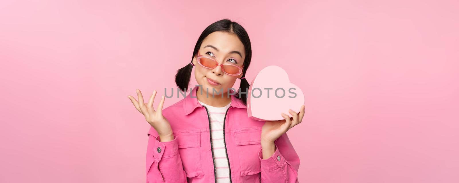 Image of stylish asian girlfriend in sunglasses, guessing whats inside gift box, heart-shaped present, standing over pink background by Benzoix