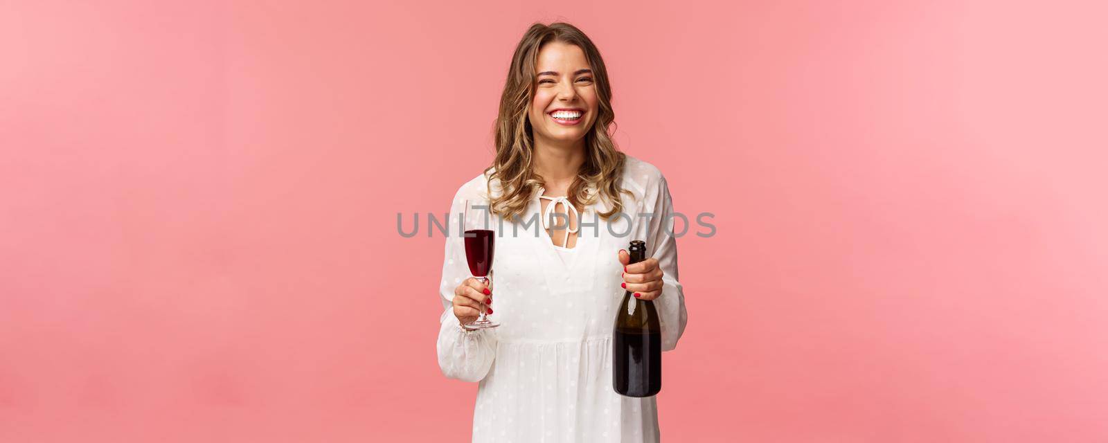 Holidays, spring and party concept. Portrait of happy and carefree european blond female celebrating in white dress, holding bottle champagne or wine, drinking from glass and laughing.