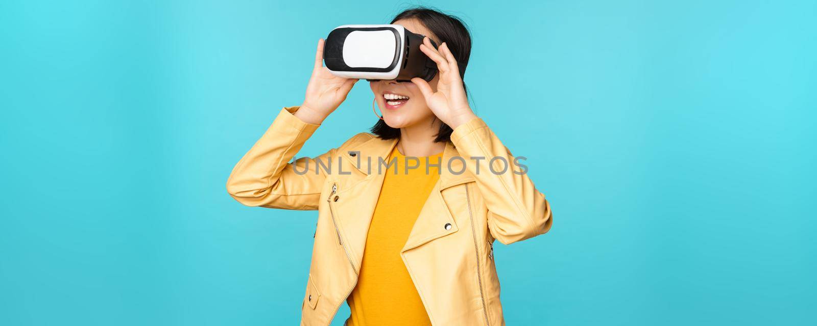 Portrait of happy asian female model using VR headset, smiling and laughing in virtual reality glasses, standing over blue background.