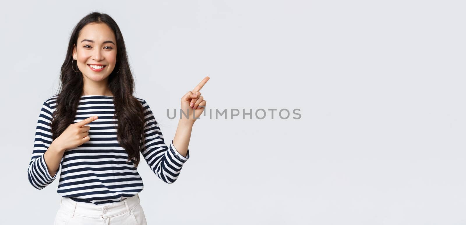 Lifestyle, beauty and fashion, people emotions concept. Good-looking young female in striped shirt pointing fingers right, inviting customers, advertise special discount, smiling at camera happy by Benzoix