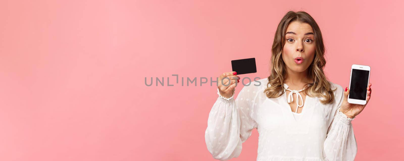 Finance, shopping and technology concept. Close-up portrait of excited blond cute girl in white dress, fold lips amused look camera as showing credit card and mobile phone, pink background by Benzoix