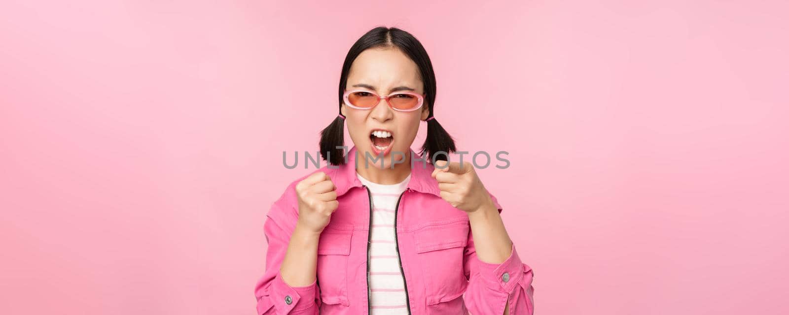 Image of angry, pissed off korean adult female model, shaking fists and shouting, screaming outraged, standing over pink background.