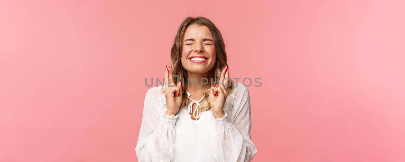 Portrait of excited hopeful blond girl making wish crossed fingers for good luck, close eyes and smiling putting all effort into pray, pleading for dream come true, anticipating over pink background.