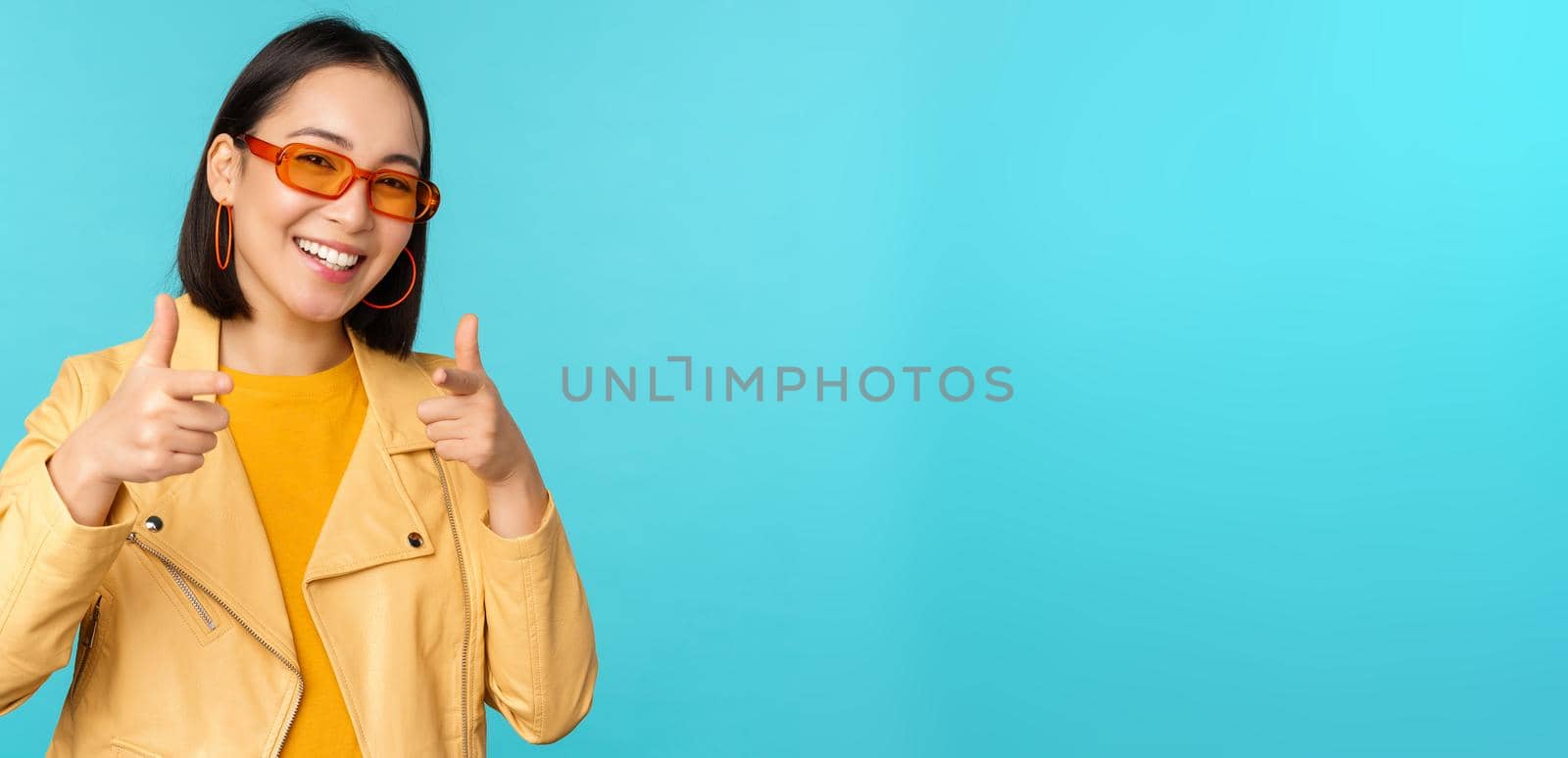 Happy young stylish chinese girl in sunglasses, points fingers at camera with pleased smile, choosing you, congratulating, standing over blue background.