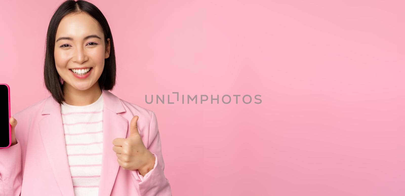 Satisfied smiling asian businesswoman recommending mobile phone app, website company on smartphone, showing screen and thumbs up, pink background by Benzoix