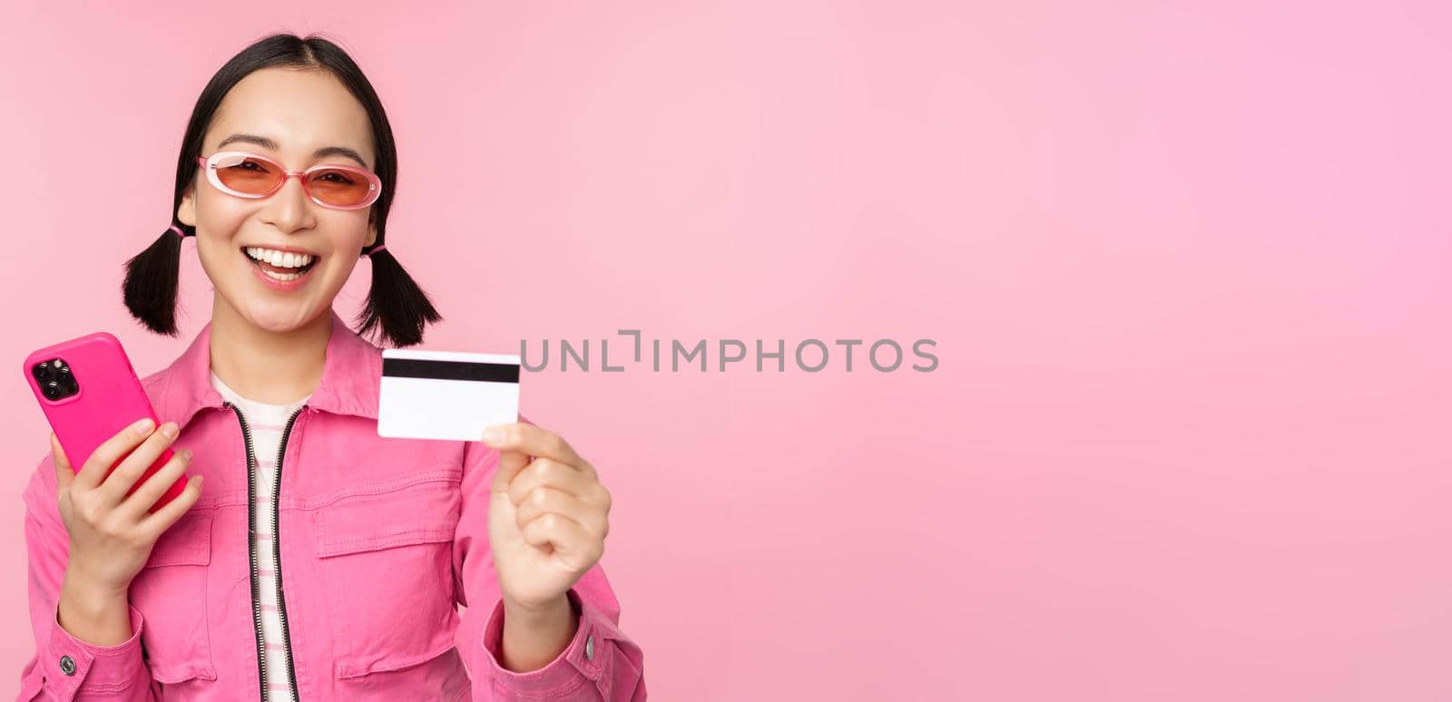 Modern beautiful asian girl, laughing and smiling with mobile phone, credit card, shopping online, paying with smartphone, standing over pink background by Benzoix