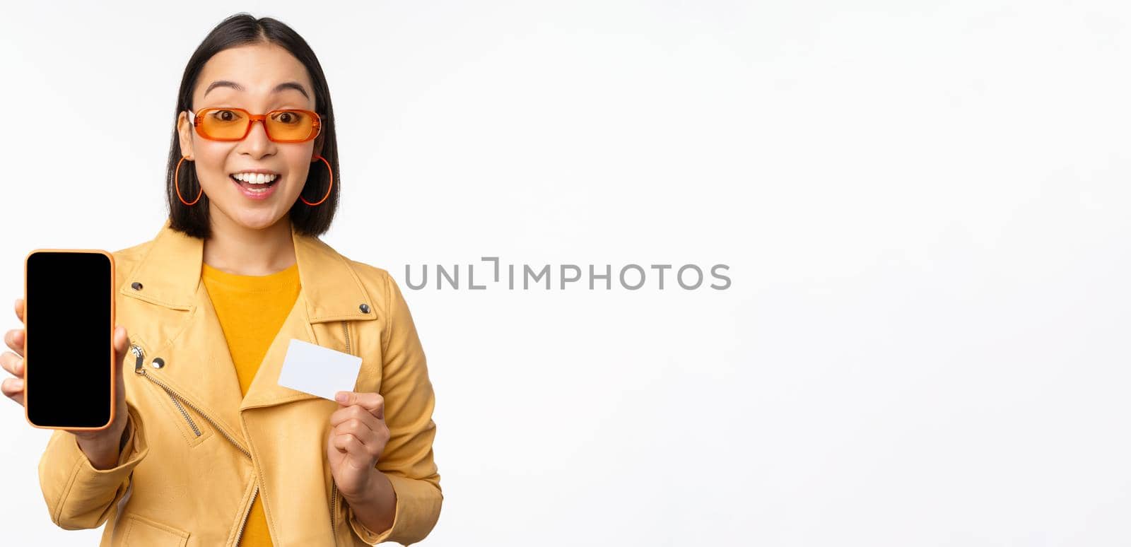Online shopping and people concept. Stylish asian woman showing mobile phone screen and credit card, smartphone application, standing over white background. Copy space