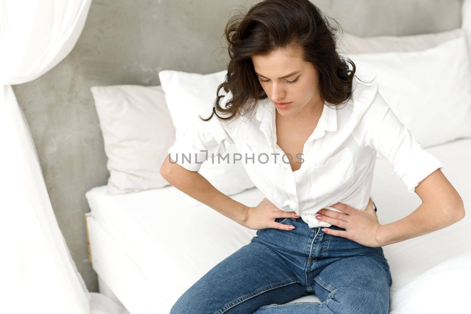Young sick woman presses her hands to her lower abdomen. Medical or gynecological problems, healthcare concept. Young woman suffering from abdominal pain, menses while sitting on bed at home.