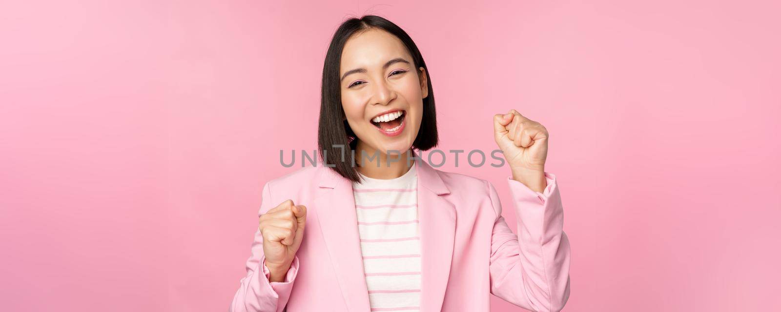 Enthusiastic saleswoman, asian corporate woman say yes, achieve goal and celebrating, triumphing, looking with rejoice and smiling, standing over pink background.