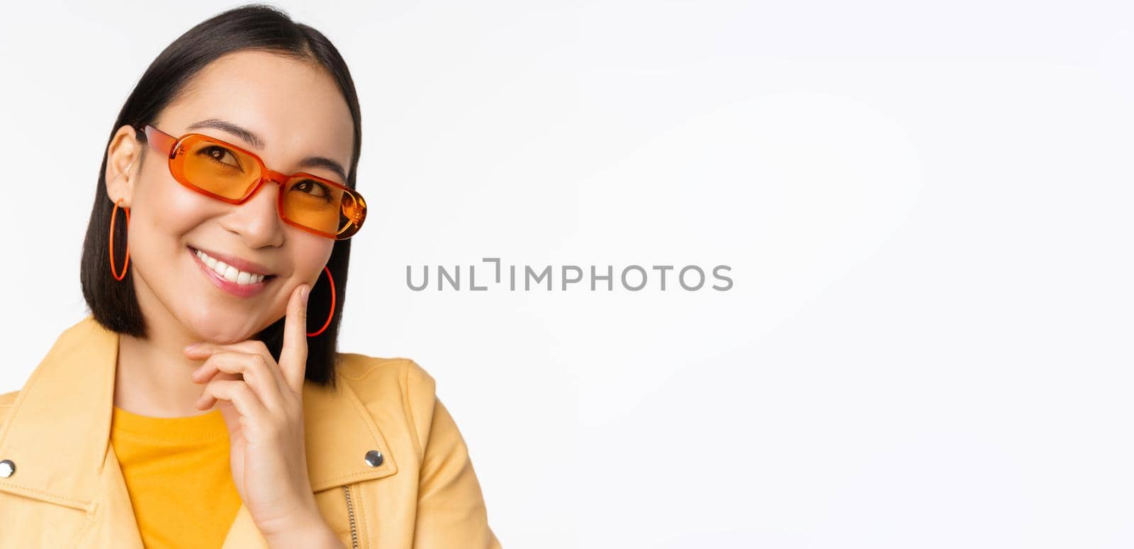 Close up portrait of asian woman thinking, wearing sunglasses and smiling, looking up thoughtful, standing over white studio background. Copy space