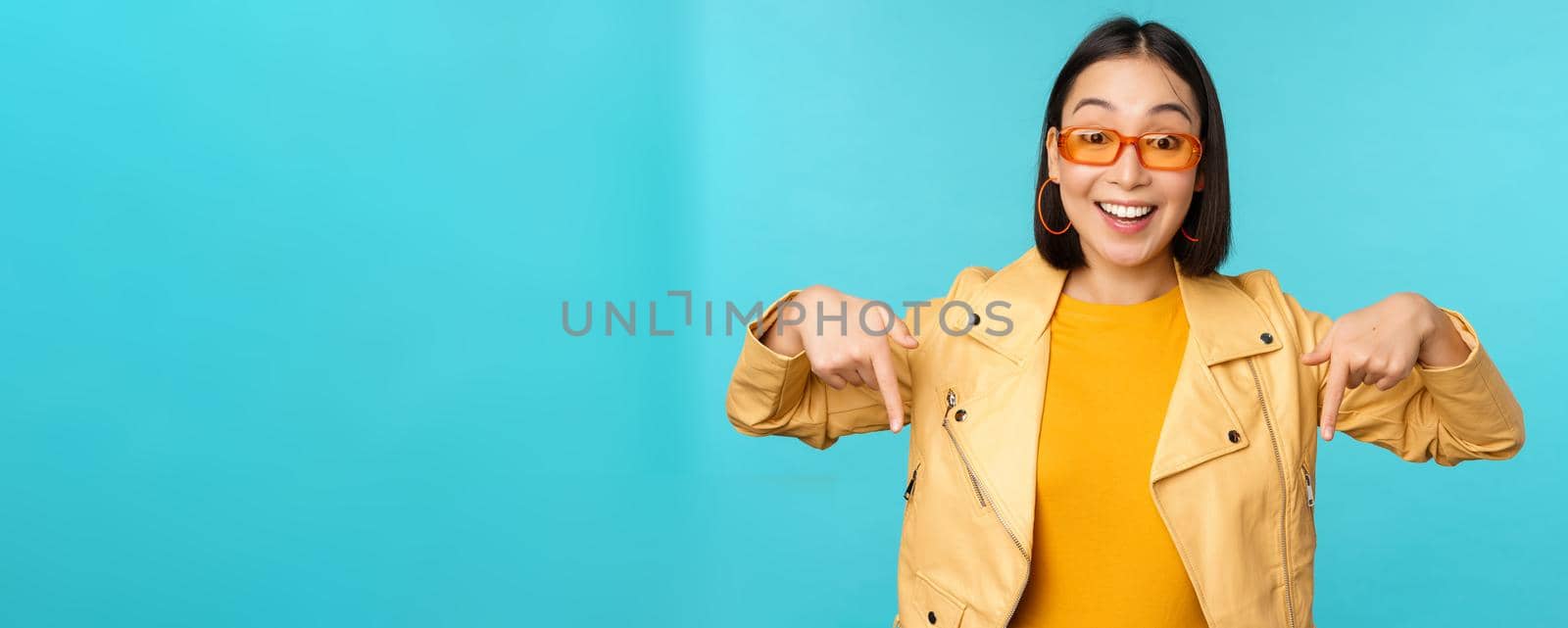 Portrait of stylish asian girl, wears sunglasses, smiles and points fingers down, shows advertisement, stands over blue background.