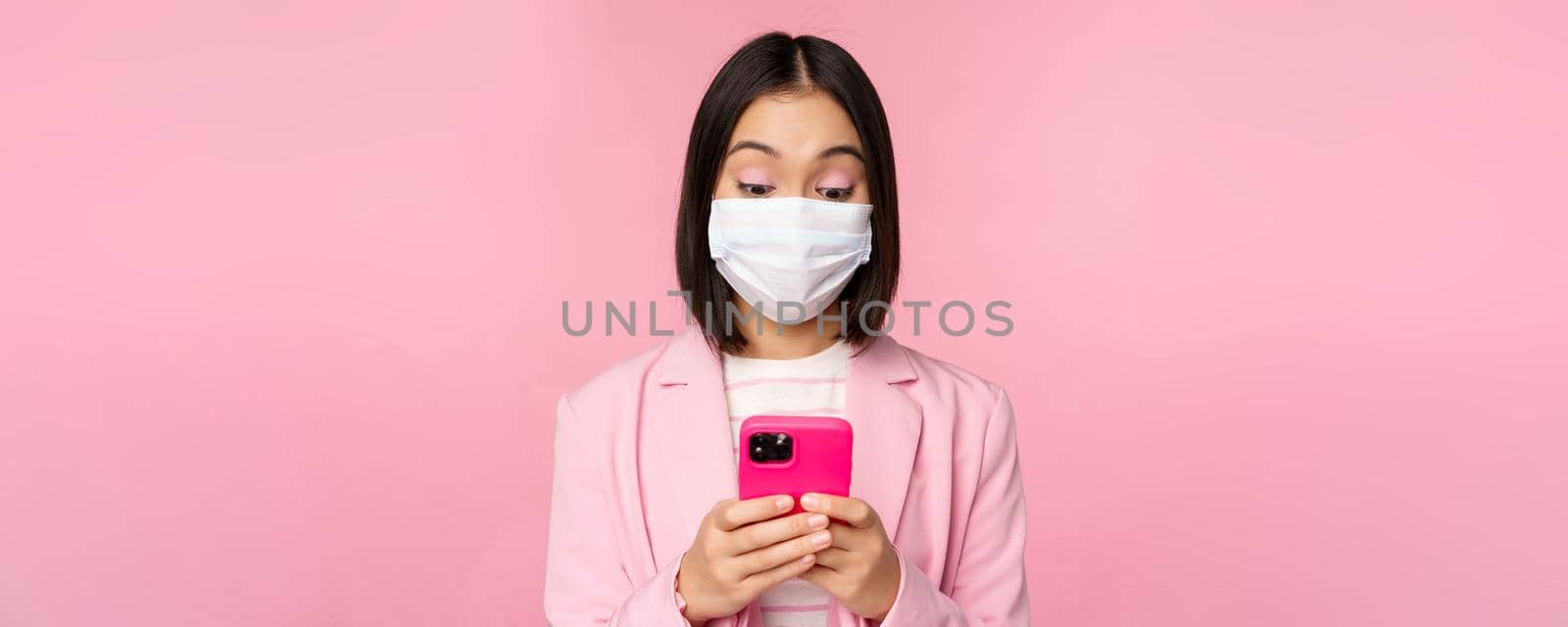 Business people and covid-19 concept. Young asian businesswoman in suit and medical face mask, using smartphone app, standing over pink background by Benzoix