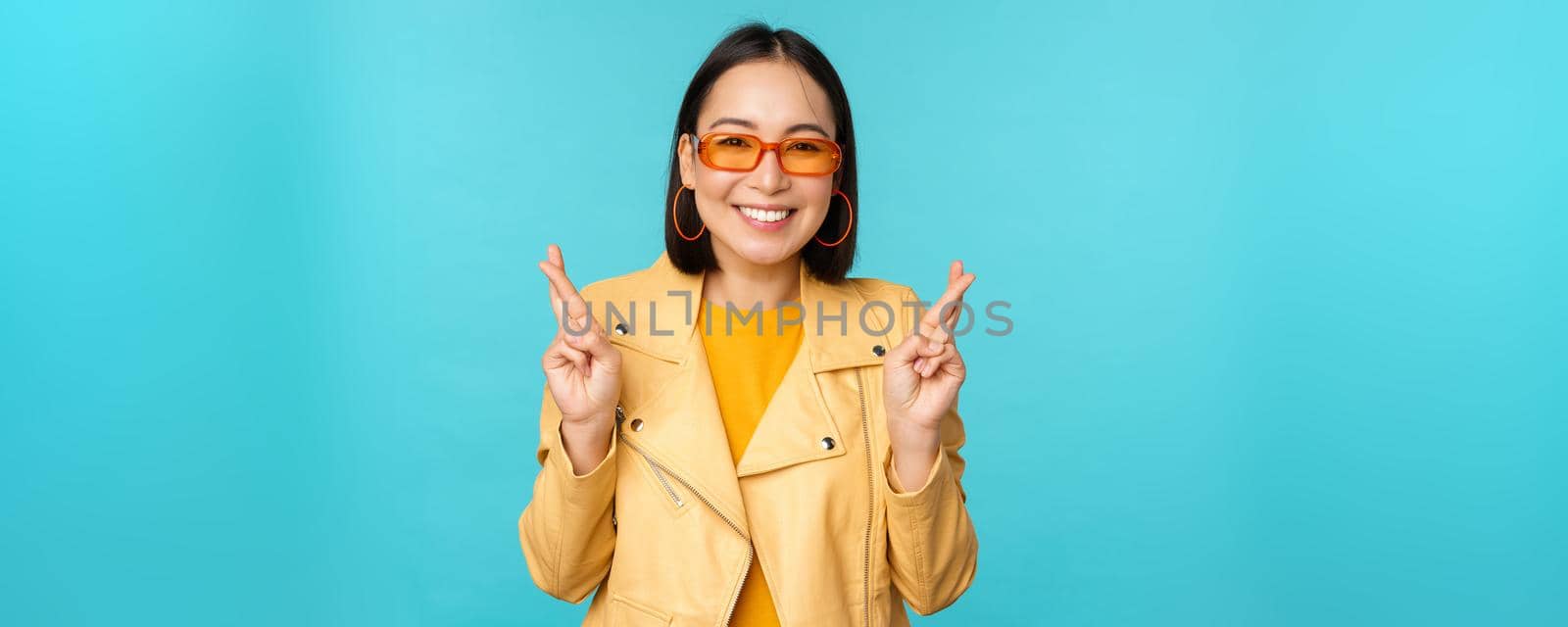 Smiling beautiful asian woman wishing, cross fingers for good luck and looking hopeful, standing over blue background. Copy space