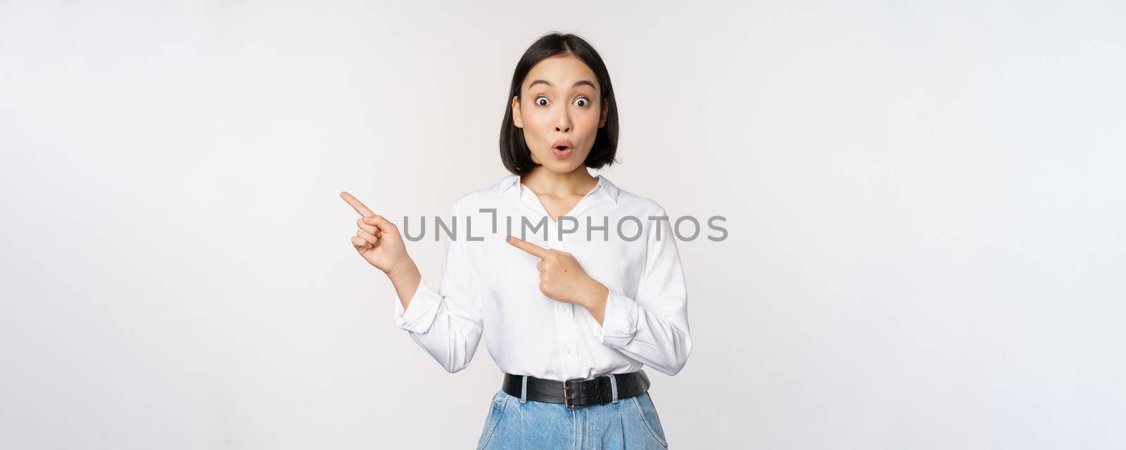 Portrait of excited young asian woman, office lady pointing fingers left at discount, showing sale banner, standing over white background.