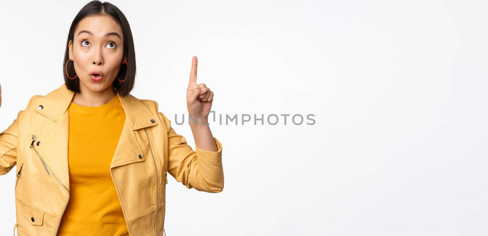 Image of smiling asian brunette woman pointing fingers up, showing advertisement with happy face, posing against white background.
