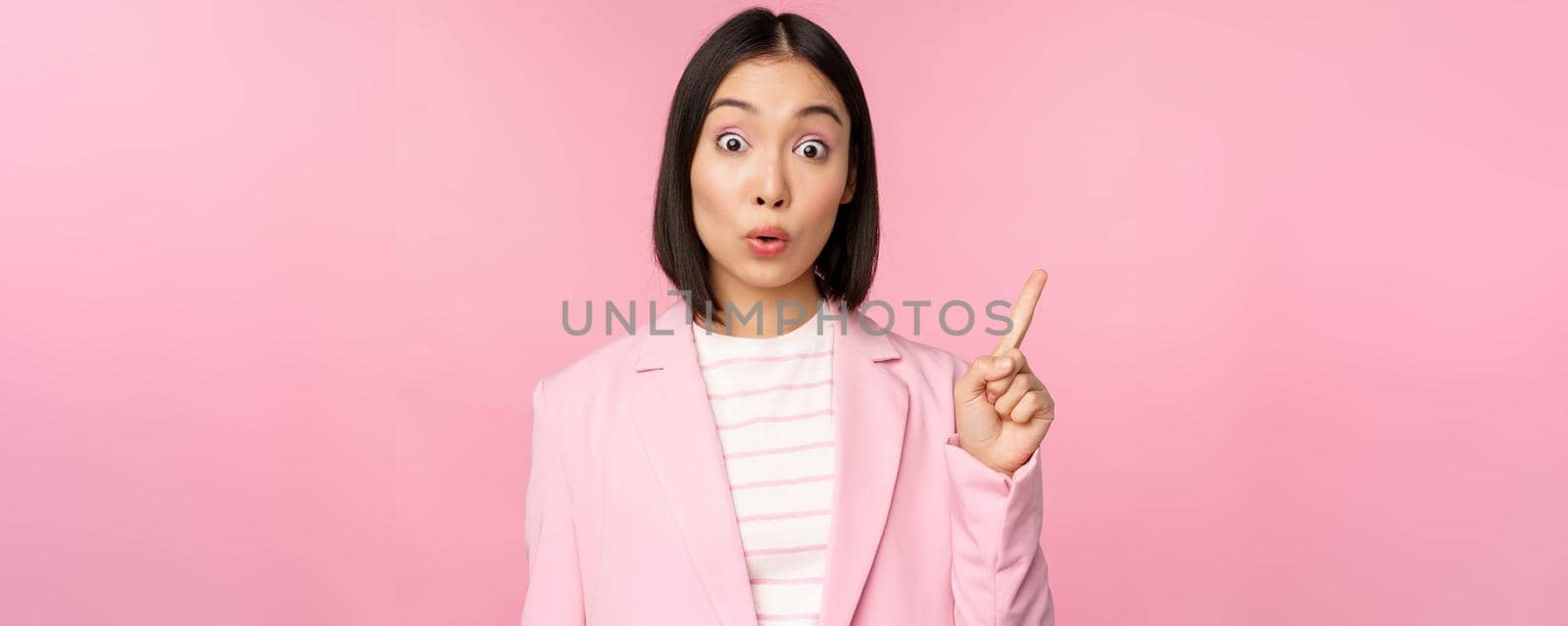 Got an idea. Young asian saleswoman, office manager raising finger, suggesting, wearing suit, posing against pink studio background.