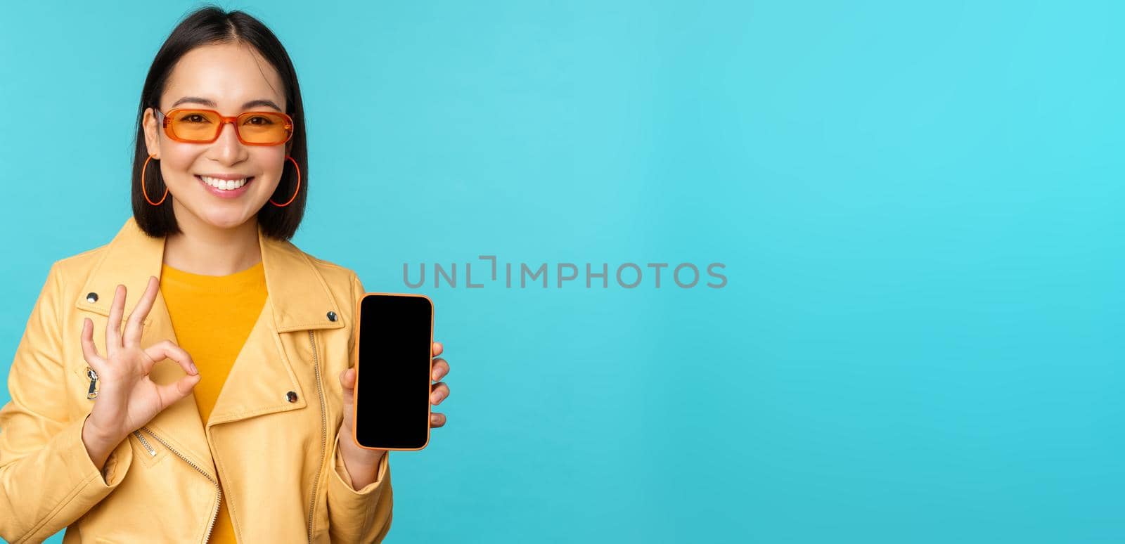 Smiling korean woman showing mobile phone app interface, smartphone application, recommending on cellphone, standing over blue background by Benzoix