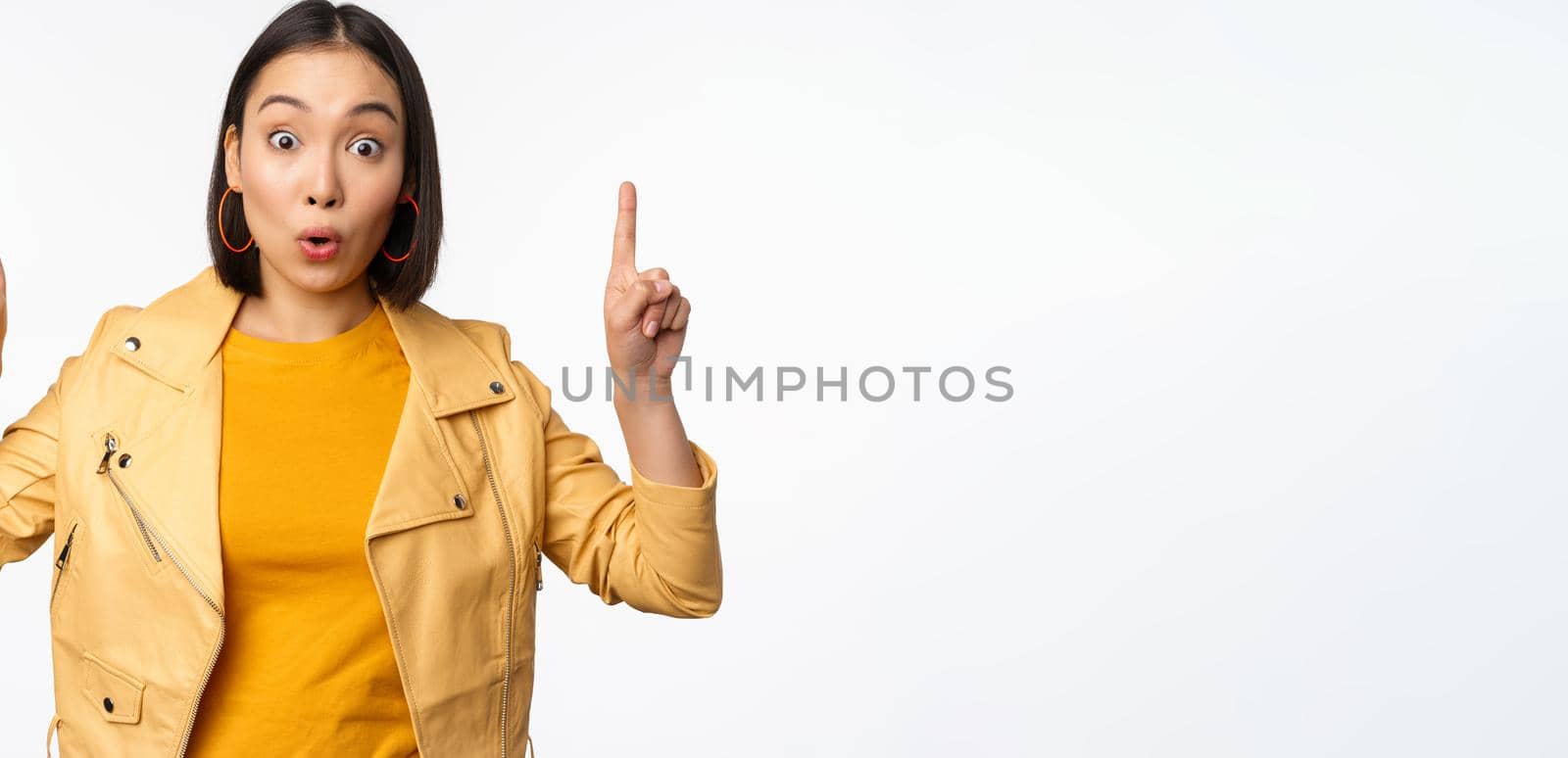 Surprised asian girl pointing fingers up, express interest, showing advertisement ahead, demonstrating promo banner on top, standing against white background.