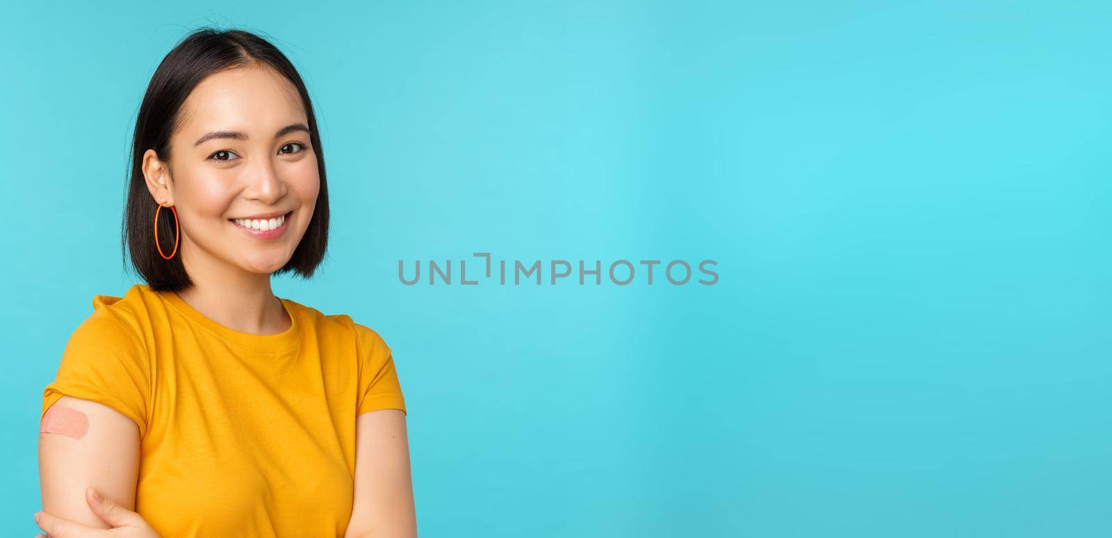 Vaccine campaign from covid-19. Young beautiful, healthy asian woman showing shoulder with bandaid, concept of vaccination, standing over blue background.