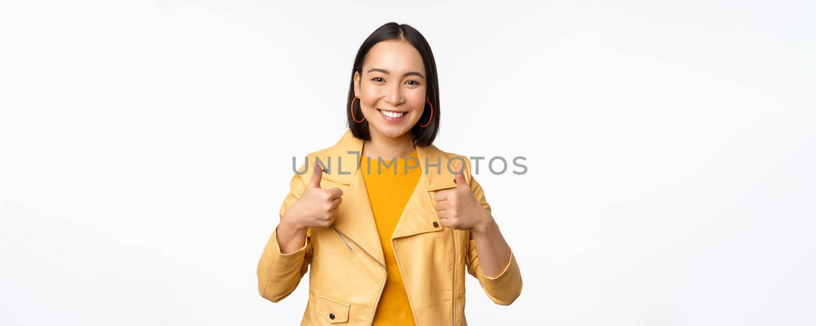 Cheerful asian woman smiling pleased, showing thumbs up in approval, standing in casual clothes over white background.