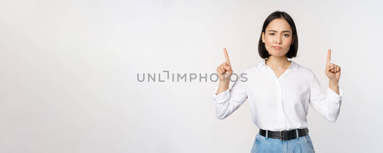 Image of skeptical asian lady grimacing, frowning upset, pointing fingers up, showing advertisement, standing over white background.