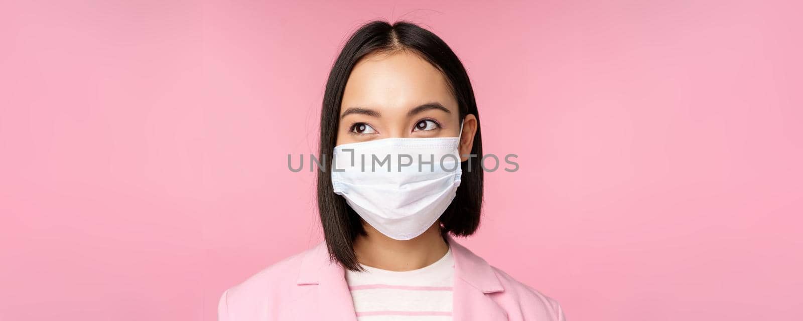 Close up portrait of japanese corporate woman in medical face mask from covid-19, looking left at logo, sale promo, standing over pink background.