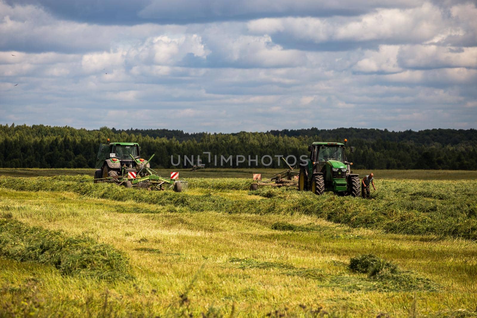 two green haymaking John Deere tractors with Krone plow on summer field before storm - telephoto shot with selective focus in Tula, Russia - July 30, 2019