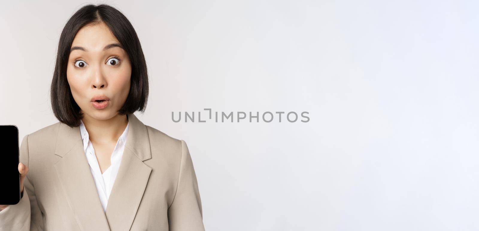 Image of asian corporate woman showing app interface, mobile phone screen, making surprised face expression, wow, standing over white background.