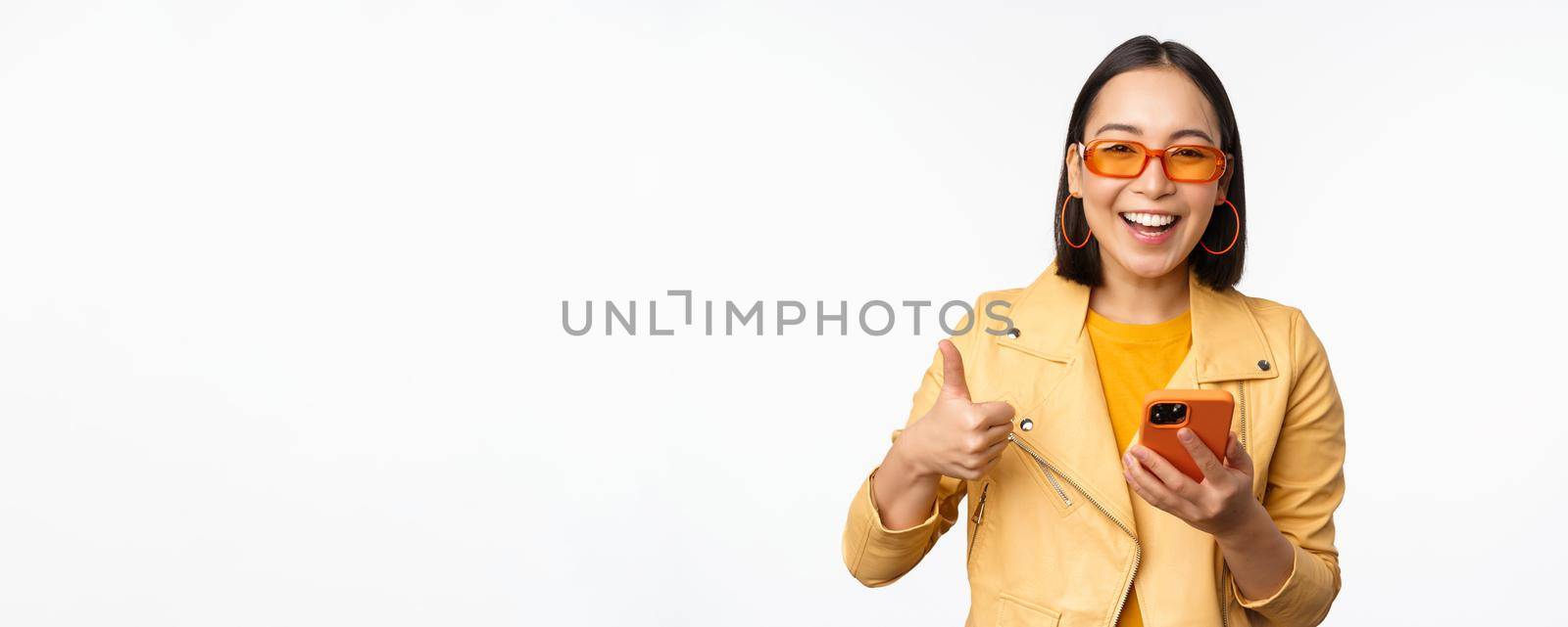 Happy stylish korean girl, wearing sunglasses, showing thumb up, holding smartphone, using mobile phone app, recommending application, online store, white background by Benzoix