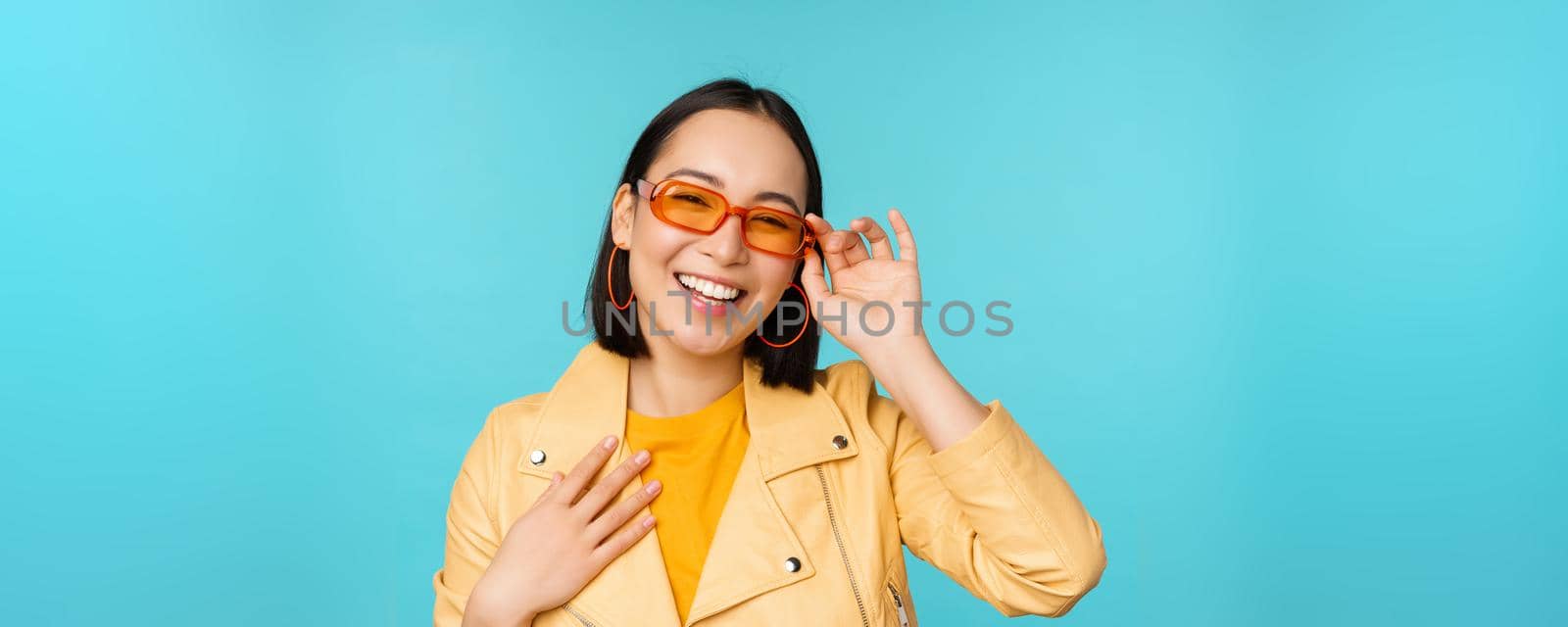 Close up portrait of stylish asian woman in sunglasses, laughing and smiling, looking happy, posing in trendy clothes over blue background by Benzoix