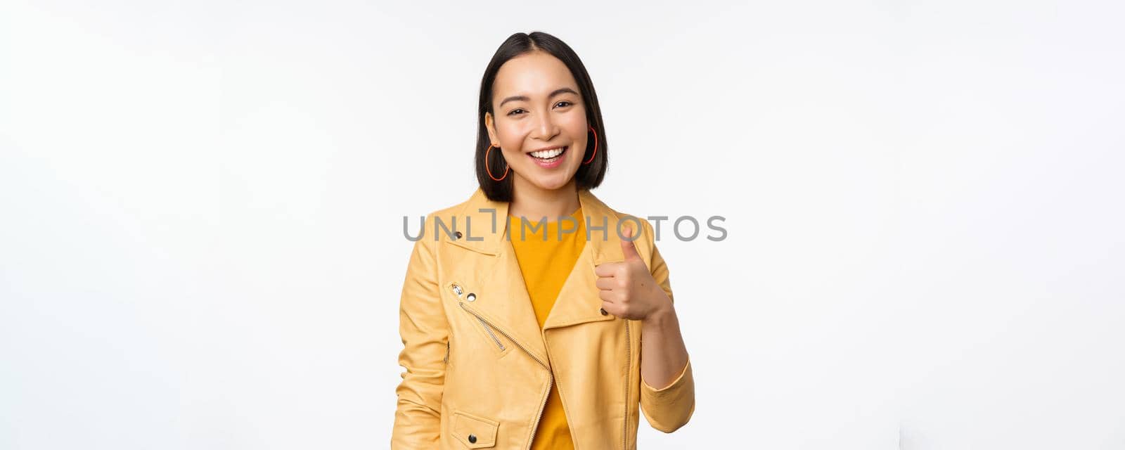 Beautiful korean girl smiling, showing thumbs up, like gesture, recommending store or company, standing satisfied against white background.