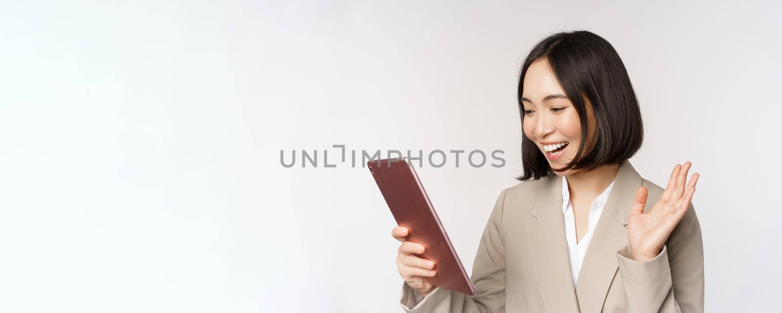 Portrait of smiling asian businesswoman video chat on digital tablet, waving hand at gadget screen, standing in suit over white background.