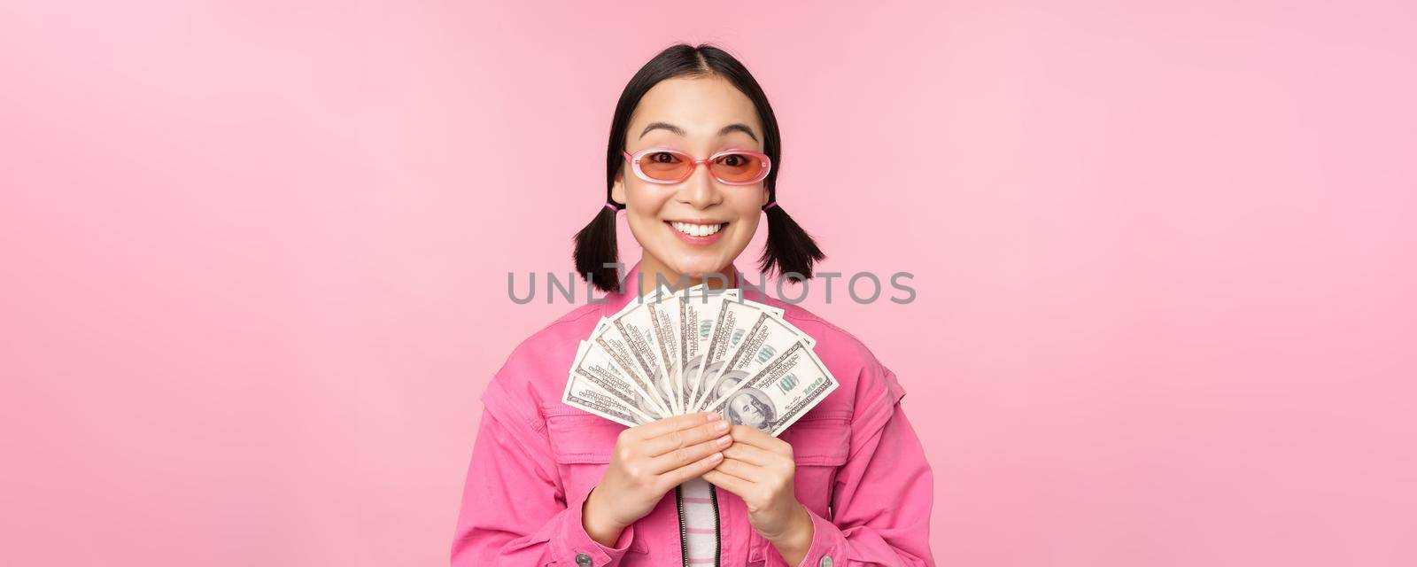 Microcredit and fast loans concept. Excited stylish korean girl, showing money, cash dollars and looking happy, standing in sunglasses over pink background.