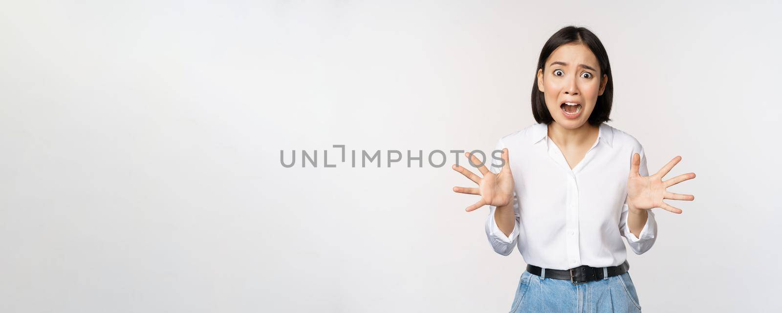 Asian woman looks at camera and screams in panic. Young korean girl looking anxious, panicking, shaking hands and shouting, standing over white background.