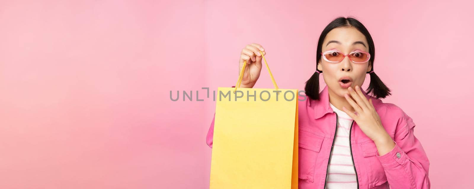 Shopping. Stylish asian girl in sunglasses, showing bag from shop and smiling, recommending sale promo in store, standing over pink background.