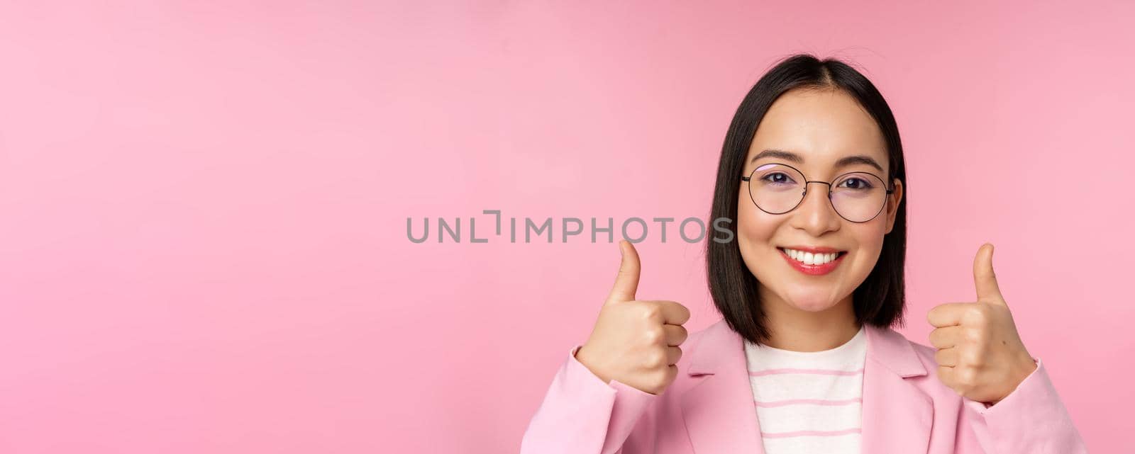 Awesome, congrats. Face of excited asian businesswoman in glasses, smiling pleased, showing thumbs up in approval, standing over pink background by Benzoix