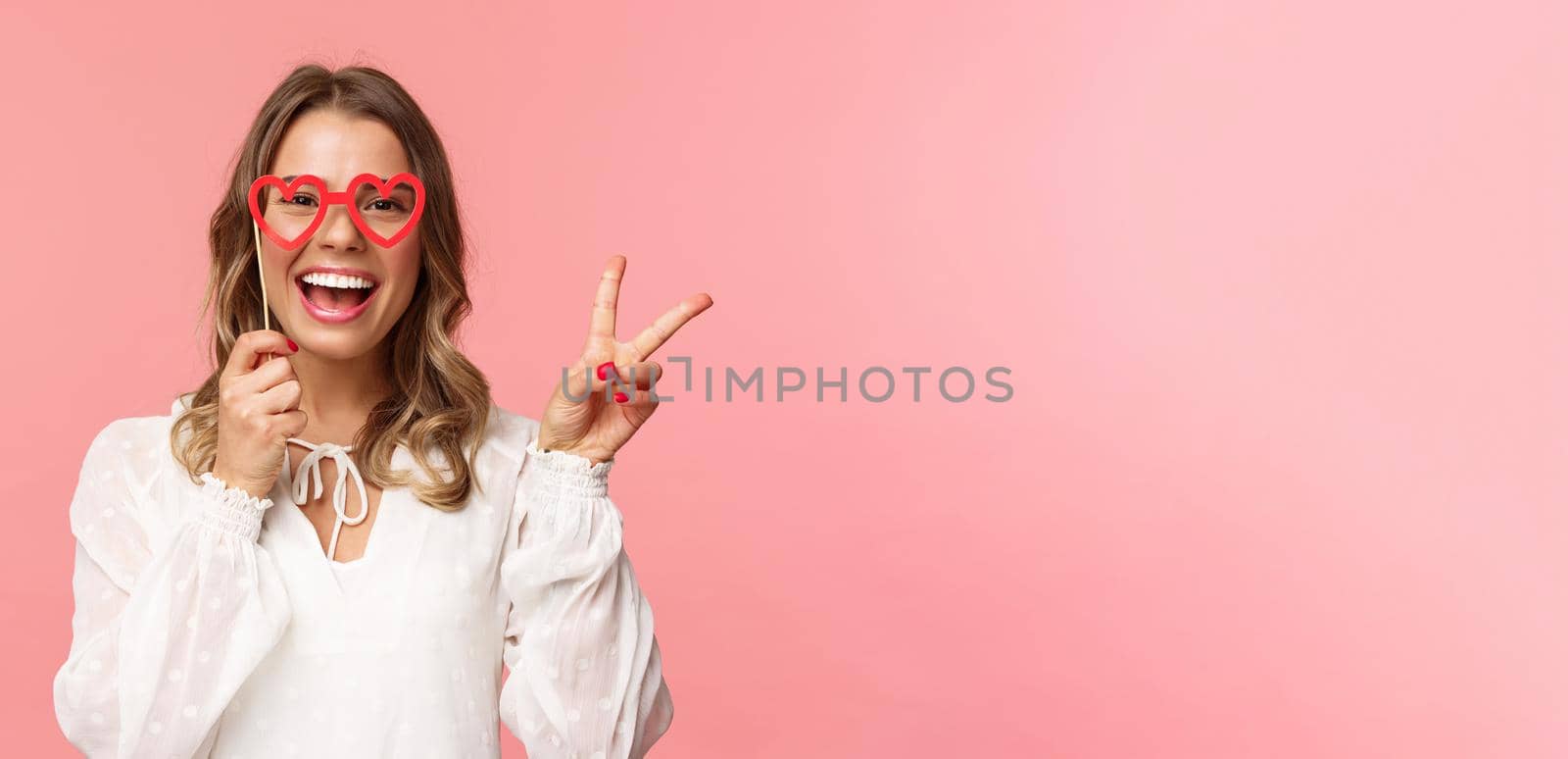 Spring, happiness and celebration concept. Close-up portrait of upbeat blond girl at party, wear white dress holding heart-shaped glasses mask over eyes and make peace sign cheerful.