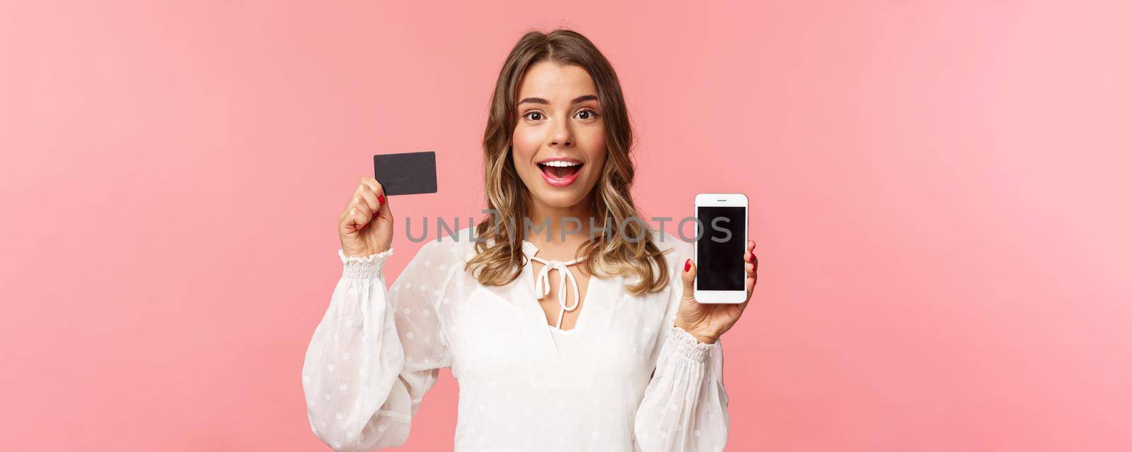 Finance, shopping and technology concept. Close-up portrait of excited blond attractive girl in white dress, showing credit card and mobile phone, advertise online store, smartphone app.