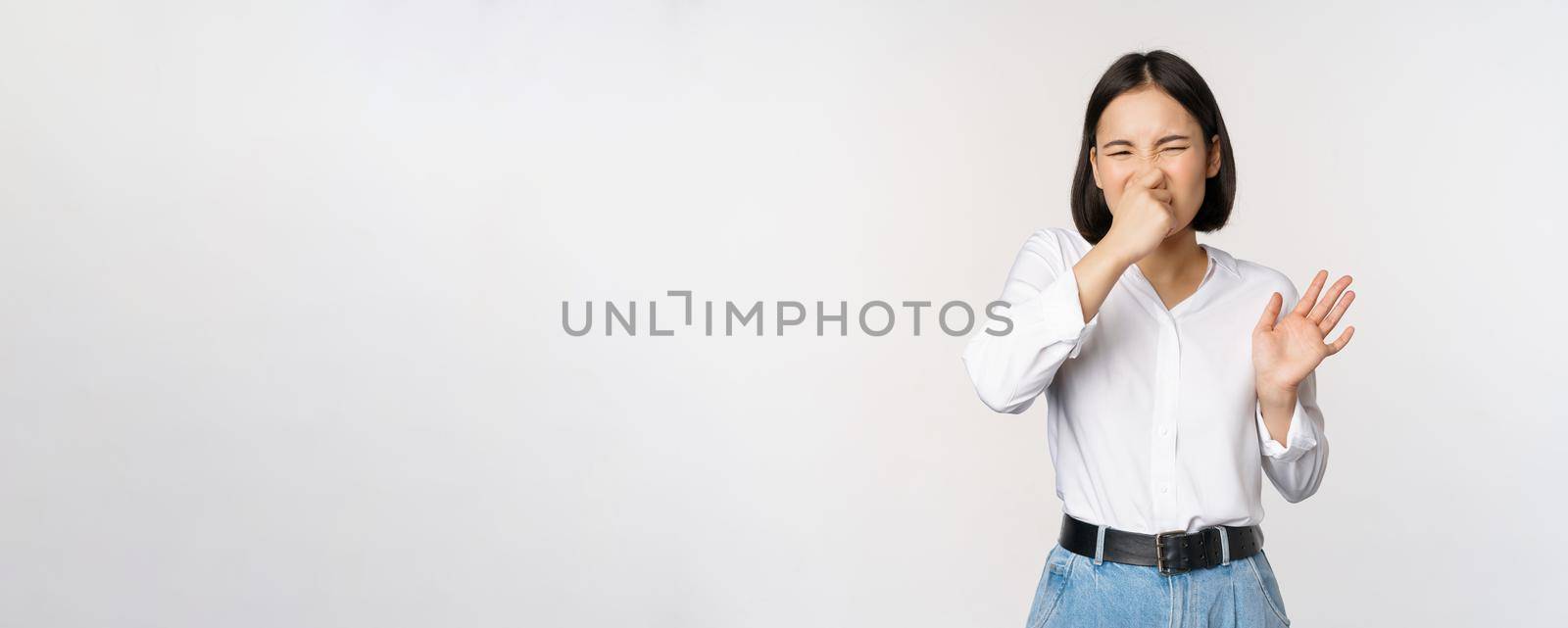 Disgusted asian girl close nose and showing rejection gesture, digusting bad smell, standing over white background.