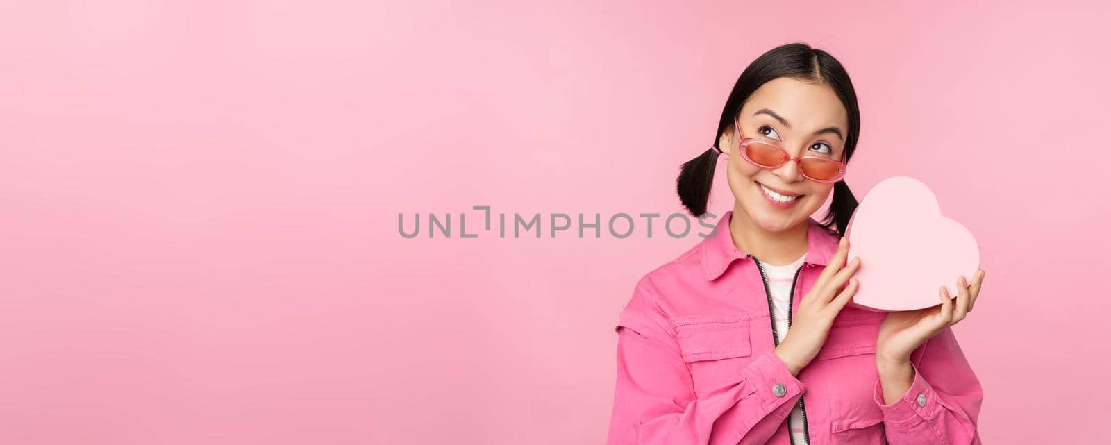 Image of stylish asian girlfriend in sunglasses, guessing whats inside gift box, heart-shaped present, standing over pink background by Benzoix