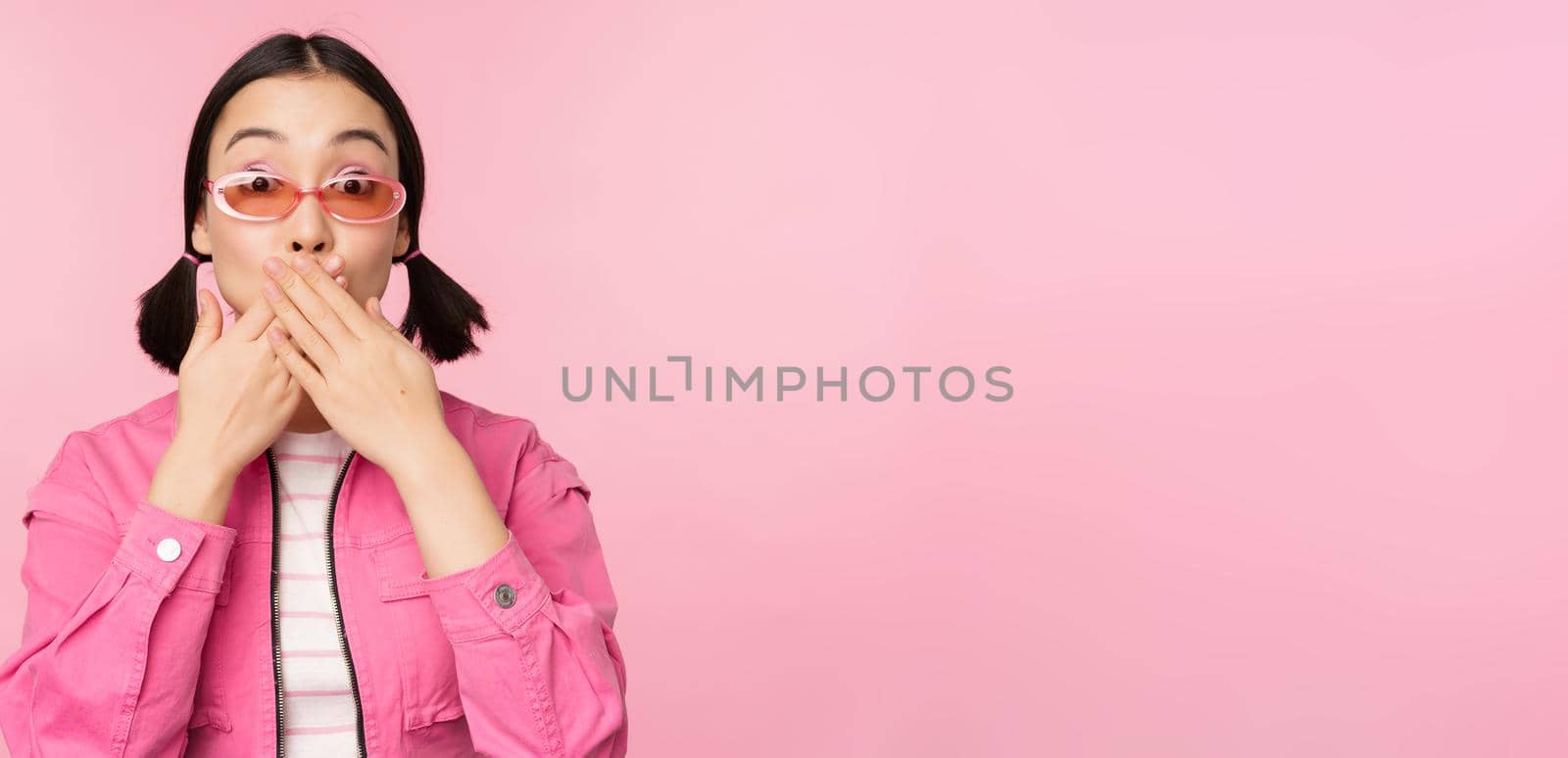 Portrait of shocked, stylish asian girl in sunglasses, closes mouth, looks with surprised face expression, stands over pink background by Benzoix