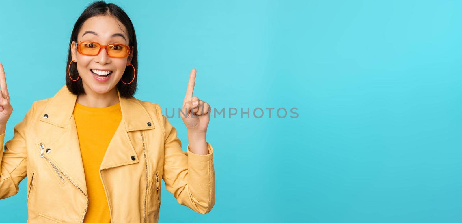 Enthusiastic asian girl in sunglasses, points fingers up, shows banner or logo on top, stands over blue background by Benzoix