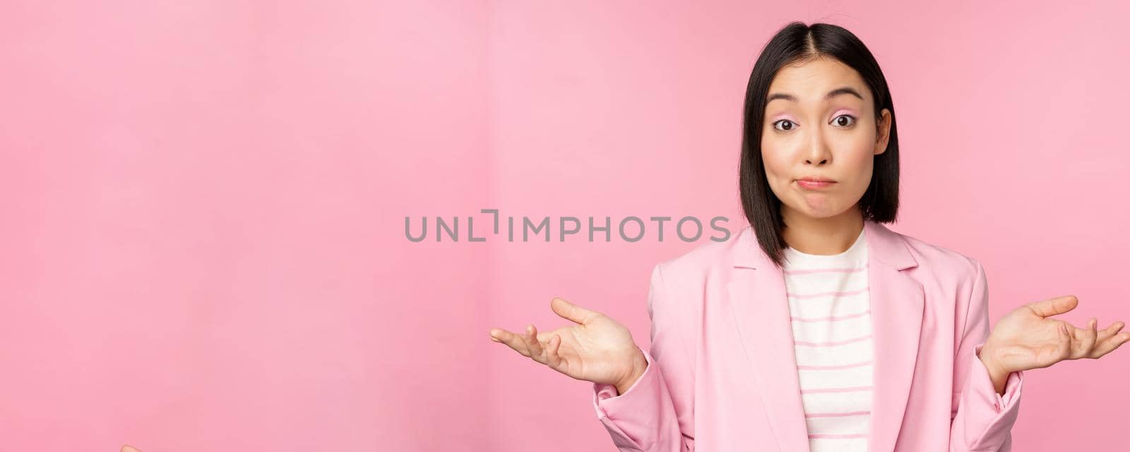 Portrait of confused asian businesswoman shrugging shoulders, looking clueless and puzzled, dont know, cant say, standing over pink background in office suit.