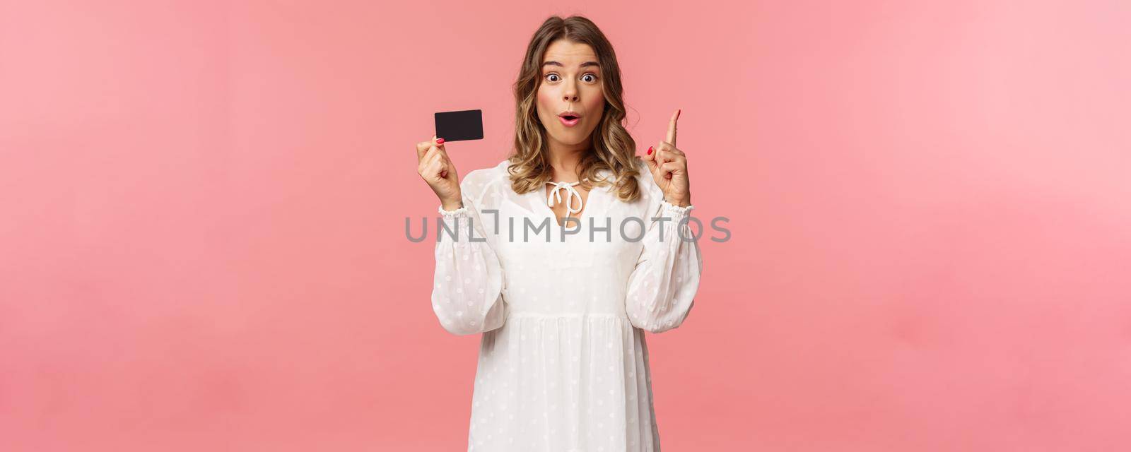 Portrait of thoughtful cute blond girl in white dress, have and excellent idea, holding credit card say wow, eureka, raise index finger have suggestion, know where find needed product by Benzoix