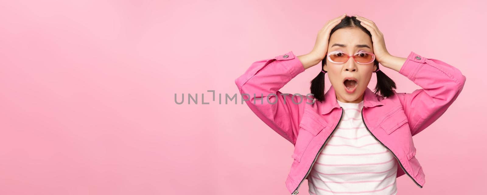 Portrait of troubled asian woman, korean girl holds hands on head and look in panic, standing distressed and shocked against pink background by Benzoix