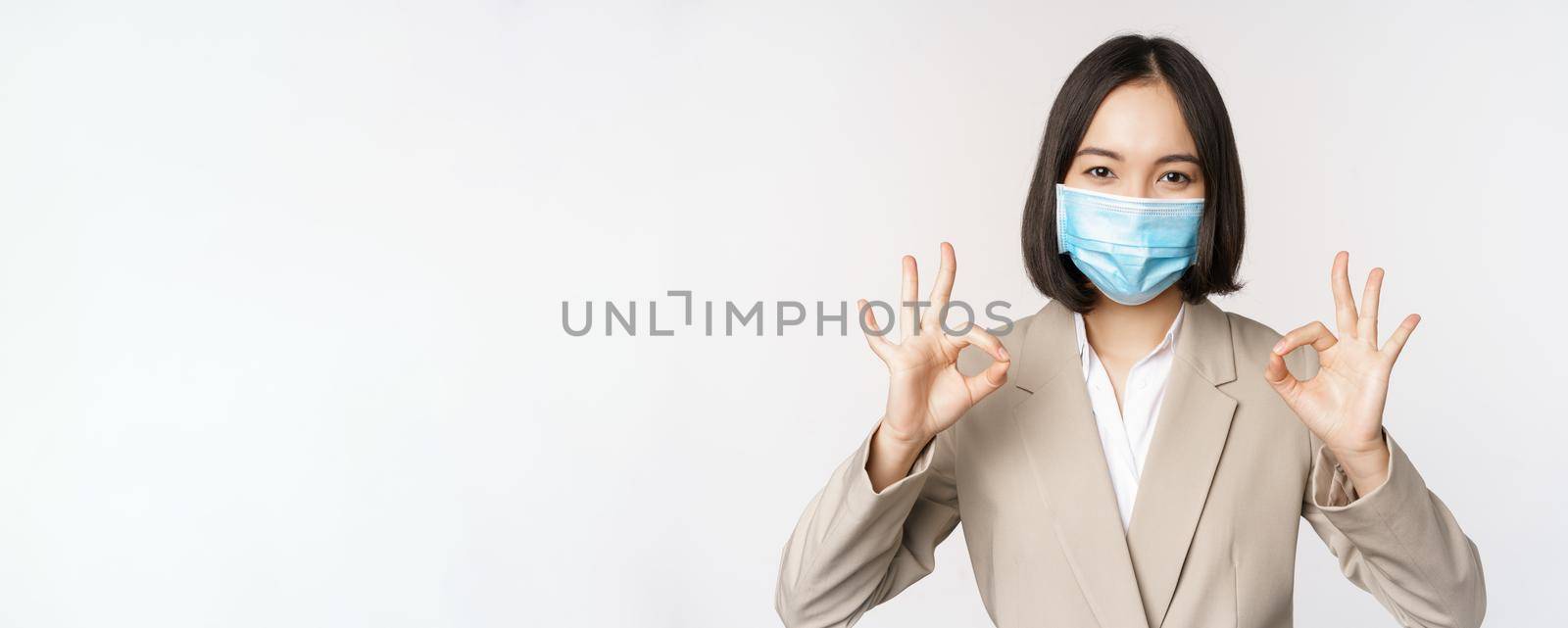 Coronavirus and workplace concept. Image of asian saleswoman, company worker in medical mask showing okay sign, smiling pleased, white background.